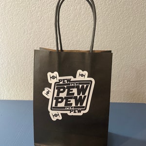 May include: A black paper gift bag with a white sticker that says "PEW PEW PEW" in bold black letters. The bag has gray rope handles.