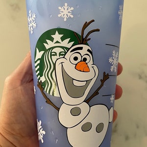 Puede incluir: Un vaso fr&iacute;o Starbucks azul claro con el personaje Olaf de la pel&iacute;cula Frozen. La taza tiene dise&ntilde;os de copos de nieve blancos y el logotipo de Starbucks. La taza es de pl&aacute;stico y tiene una tapa azul.