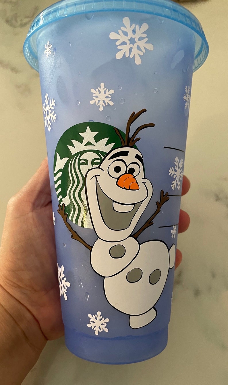 Puede incluir: Un vaso fr&iacute;o Starbucks azul claro con el personaje Olaf de la pel&iacute;cula Frozen. La taza tiene dise&ntilde;os de copos de nieve blancos y el logotipo de Starbucks. La taza es de pl&aacute;stico y tiene una tapa azul.