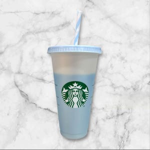 Puede incluir: Un vaso fr&iacute;o Starbucks azul claro con tapa a juego y pajita a rayas azules y blancas. El logotipo de Starbucks es verde. La taza est&aacute; llena de l&iacute;quido azul claro.