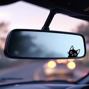 Puede incluir: Una pegatina de un gato negro asom&aacute;ndose por el espejo retrovisor de un coche. El gato tiene ojos grandes y una expresi&oacute;n curiosa. El espejo refleja la carretera, con luces borrosas en el fondo.