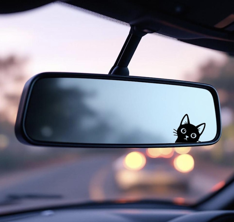 Puede incluir: Una pegatina de un gato negro asom&aacute;ndose por el espejo retrovisor de un coche. El gato tiene ojos grandes y una expresi&oacute;n curiosa. El espejo refleja la carretera, con luces borrosas en el fondo.