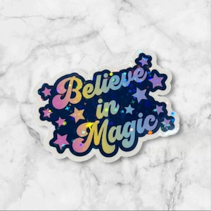 May include: A holographic sticker with the words "Believe in Magic" in a retro font. The sticker has a dark blue background with colorful stars and a rainbow gradient effect.