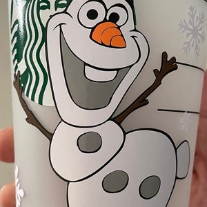 Puede incluir: Un vaso Starbucks azul claro con Olaf de Frozen sonriendo. El dise&ntilde;o incluye copos de nieve blancos y el logotipo de Starbucks. Olaf tiene una nariz de zanahoria y brazos de ramitas. Una divertida taza con tem&aacute;tica invernal.