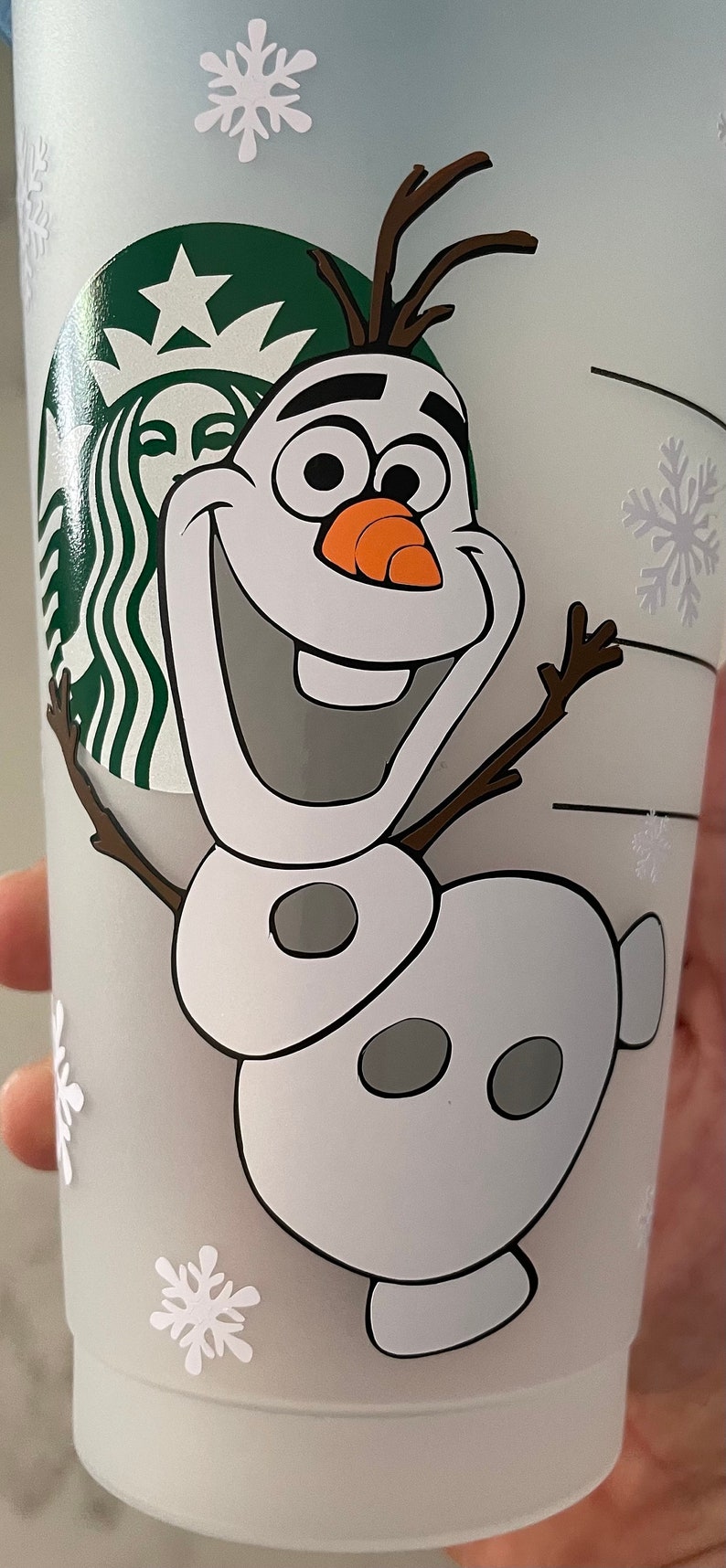 Puede incluir: Un vaso Starbucks azul claro con Olaf de Frozen sonriendo. El dise&ntilde;o incluye copos de nieve blancos y el logotipo de Starbucks. Olaf tiene una nariz de zanahoria y brazos de ramitas. Una divertida taza con tem&aacute;tica invernal.