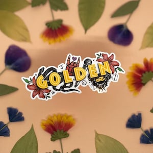 Pode incluir: Um adesivo com a palavra "GOLDEN" em letras de néon amarelas, acompanhado por uma cobra preta, um microfone, uma mão fazendo o sinal de rock and roll e flores.
