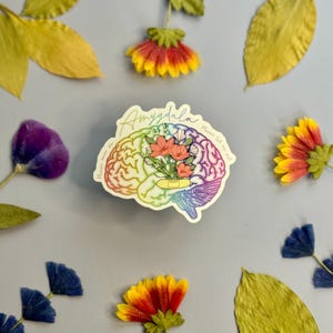 Pode incluir: Um adesivo recortado com uma ilustração de um cérebro humano com um gradiente de arco-íris, um buquê de flores rosas e um curativo adesivo bege. O texto "Amygdala" e "Please save me, please let me out" está impresso em cursivo.