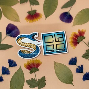 May include: A blue snake sticker with a yellow stripe wraps around a blue and yellow window sticker with the text "CLA" inside. The stickers are on a light brown background with dried flowers.