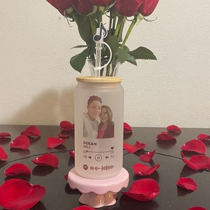 May include: A frosted glass tumbler with a photo of a couple and the song title "Ocean" by Karol G printed on it. The tumbler is on a pink cake stand surrounded by red rose petals.