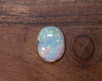 May include: A white opal gemstone with iridescent blue, green, and orange flashes. The opal is oval shaped and is set against a brown wooden background.
