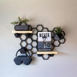 May include: Black honeycomb-shaped wall shelf with two wooden shelves. The shelf holds a bonsai tree, a book titled "Never Finished" by David Goggins, a geometric planter with a succulent, and a pair of black headphones.