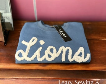 May include: A blue sweatshirt with a white fluffy embroidered word "lions" on the front. The sweatshirt is folded and has a black tag with the word "Leary" on it.