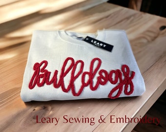 May include: A white sweatshirt with a red embroidered "Bulldog" design. The sweatshirt is folded and laid on a wooden surface. The text "Leary Sewing & Embroidery" is visible at the bottom of the image.
