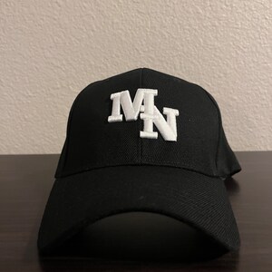 以下が含まれることがあります： 前面に白い刺繍で「MN」のロゴが入った黒い野球帽
