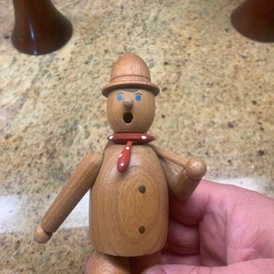 Puede incluir: Una figurita de madera de una persona que lleva un sombrero marrón, una bufanda roja con lunares blancos y una camisa a rayas azules y blancas. La figurita tiene una cara pintada con ojos azules y una boca roja.