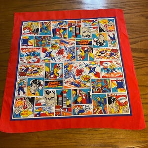 May include: A square red bandana with a comic book-style print. The design features colorful panels with action scenes, characters, and speech bubbles. The border is a solid red, and the interior has a blue border.