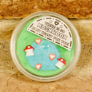 Könnte beinhalten: Ein durchsichtiger Kunststoffbehälter gefüllt mit grünem und blauem Schleim, verziert mit pilz- und herzförmigen Verzierungen. Der Behälter hat ein Etikett mit der Aufschrift "THERE IS SO MUSHROOM IN MY HEART FOR YOU!"