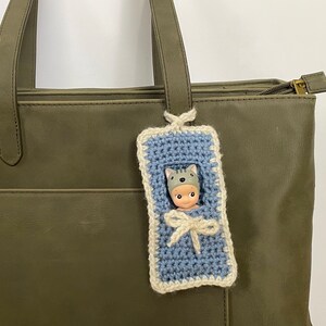 May include: A blue crocheted baby sleeping bag with a white trim, hanging from a green tote bag. A small doll with a gray cat hat is inside the sleeping bag.