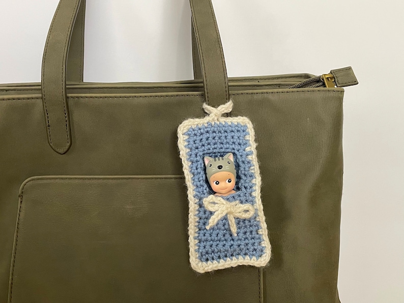 May include: A blue crocheted baby sleeping bag with a white trim, hanging from a green tote bag. A small doll with a gray cat hat is inside the sleeping bag.