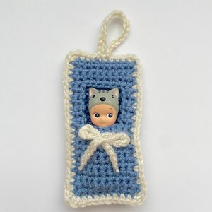 May include: A crocheted baby sleeping bag with a white trim and a doll wearing a gray cat hat inside. The sleeping bag has a white ribbon tied at the top.