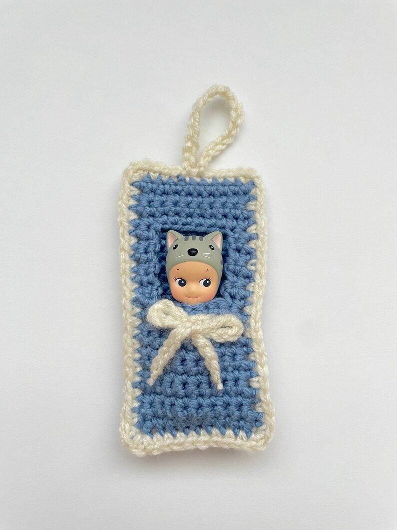 May include: A crocheted baby sleeping bag with a white trim and a doll wearing a gray cat hat inside. The sleeping bag has a white ribbon tied at the top.
