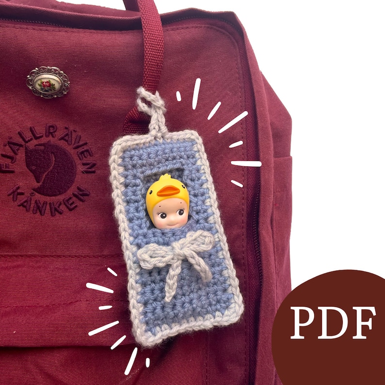 May include: A blue and white crocheted baby carrier with a yellow duck-shaped toy inside. The carrier is attached to a red backpack with the Fj&auml;llr&auml;ven K&aring;nken logo.