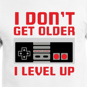 Puede incluir: Camiseta blanca con un gráfico rojo que dice "No envejezco, subo de nivel" con un gráfico de mando de videojuego gris y negro.