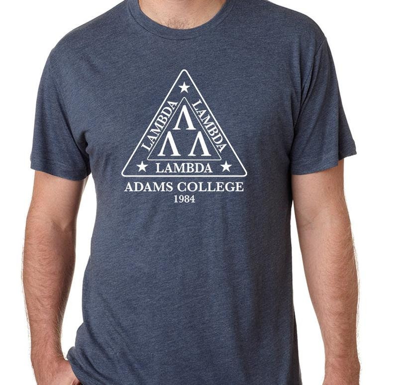 Może przedstawiać: Koszulka w kolorze granatowym z białym tr&oacute;jkątnym logo. Logo zawiera napis "Lambda" i grecką literę "Lambda" trzy razy. Pod tr&oacute;jkątem znajduje się napis "Adams College 1984".
