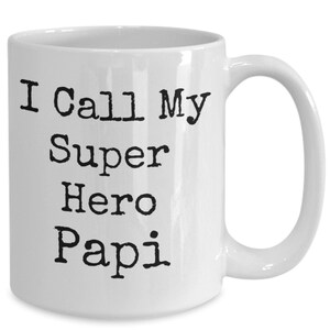 May include: White ceramic mug with the text "I Call My Super Hero Papi" in black, bold, typewriter-style font. The mug has a curved handle and is suitable for hot or cold drinks.