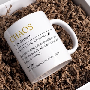 Könnte beinhalten: Eine weiße Keramiktasse mit einem schwarzen und goldenen Design. Die Tasse zeigt das Wort "CHAOS" in goldenen Buchstaben und die Definition von "Koordinator" in schwarzem Text. Die Definition beinhaltet die phonetische Aussprache des Wortes und eine Beschreibung eines "Koordinators" als jemanden, der Probleme auf unerwartete Weise löst.