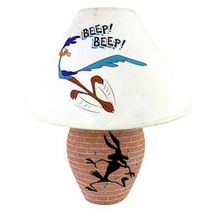 May include: A novelty lamp featuring a white lampshade with a cartoon Road Runner and the words "Beep! Beep!". The base is brick-patterned with a silhouette of Wile E. Coyote. The lamp is a fun, decorative item.