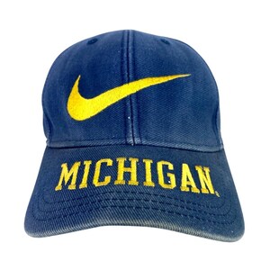 Puede incluir: Gorra de béisbol azul marino con el logotipo de Nike en amarillo y la palabra "MICHIGAN" bordada en amarillo. La gorra está hecha de tela resistente y tiene una visera curva.