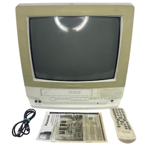 Könnte beinhalten: Ein beigefarbener und weißer Panasonic Omnivision VHS-Kombinationsfernseher mit integriertem Videorekorder und UKW-Radio. Enthält ein schwarzes Netzkabel, eine Bedienungsanleitung für die Modelle PV-C1322/PV-C1332W und eine passende beige Fernbedienung.