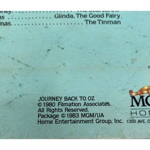 Puede incluir: Una caja de cinta VHS azul con el t&iacute;tulo "Journey Back to Oz" e informaci&oacute;n de copyright para Filmation Associates y MGM/UA Home Entertainment Group, Inc. La caja presenta un dise&ntilde;o de tira de pel&iacute;cula con el logotipo de MGM.