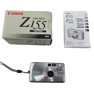 May include: A silver Canon Sure Shot Z155 Caption point-and-shoot camera with a wrist strap, accompanied by its original cardboard box labeled with the model name and a printed instruction manual featuring an image of the camera on the cover.
