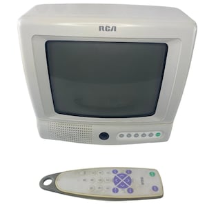 Puede incluir: Un televisor RCA blanco con pantalla oscura y mando a distancia a juego. El televisor tiene un altavoz, un botón de encendido y varios botones de control. El mando tiene botones morados y un lazo gris.