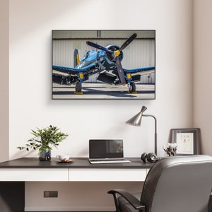 May include: A framed print of a vintage blue and yellow airplane with a large propeller. The aircraft is parked in front of a hangar. The image is displayed on a white wall above a desk with a laptop and accessories.