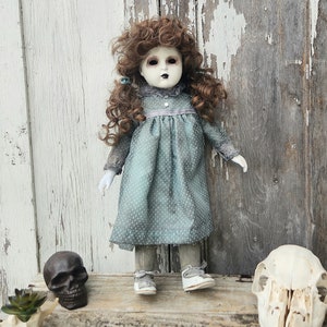 May include: A porcelain doll with white painted skin, brown curly hair, and blue eyes. The doll is wearing a blue and white polka dot dress, white shoes, and is standing in front of a weathered wooden background.