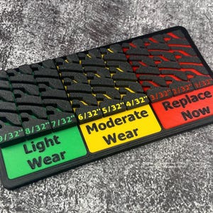 Pode incluir: Um medidor de profundidade do piso de pneus preto e multicolorido. O medidor tem seções rotuladas como "Desgaste leve", "Desgaste moderado" e "Substituir agora". Medidas em polegadas.