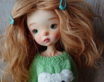 May include: A ball-jointed doll with wavy ginger hair, two light blue hair clips, and green eyes. The doll wears a light green knitted sweater featuring a textured white bear face appliqué with black eyes and a black nose on the front.