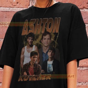 May include: Black t-shirt with a vintage-style graphic of Ashton Kutcher. The graphic features four images of Ashton Kutcher in different poses, with the text "Ashton Kutcher" in a bold, orange font.