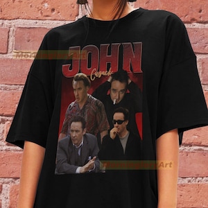 May include: Black t-shirt with a vintage-style graphic of John Cusack's face repeated four times. The text "JOHN Cusack" is printed in red and white letters above the images.