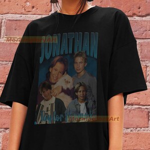 May include: Black t-shirt featuring a vintage-style graphic of four boys and a cat. The graphic includes the text "JONATHAN" in large, bold lettering.