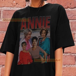 May include: Black t-shirt with a vintage-style graphic of the cast of the movie "Annie". The graphic features four women in a car, with the title "Annie" in red letters above them.