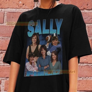 May include: Black t-shirt with a vintage-style graphic of Sally Field, featuring a collage of images from her career. The text "SALLY FIELD" is printed in blue and white.