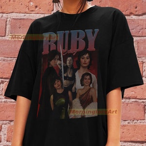 May include: Black t-shirt with a graphic print of a collage of images of women with the text "RUBY CRUZ" in a red and pink gradient.