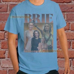 May include: A light blue t-shirt with a collage of images of Brie Larson. The text "BRIE" is in large, bold, brown letters. The text "Larson" is in smaller, brown letters. The text "MorninggloryArt" appears twice on the shirt.
