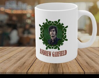May include: White ceramic coffee mug with a green holly wreath design featuring a photo of Andrew Garfield. The text "Andrew Garfield" is printed in brown below the wreath.