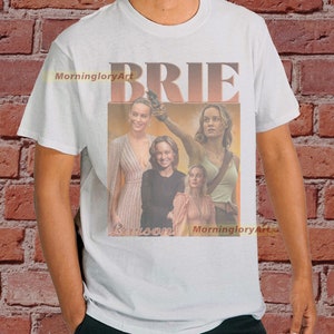 May include: White t-shirt with a collage of images of Brie Larson. The images are in a retro style with a faded, vintage look. The text "BRIE" is printed in large, bold letters across the top of the shirt.