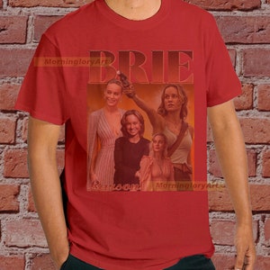 May include: Red t-shirt with a collage of images of Brie Larson, with the word "BRIE" in large, bold, white letters above the images. The text "Larson" is below the images.